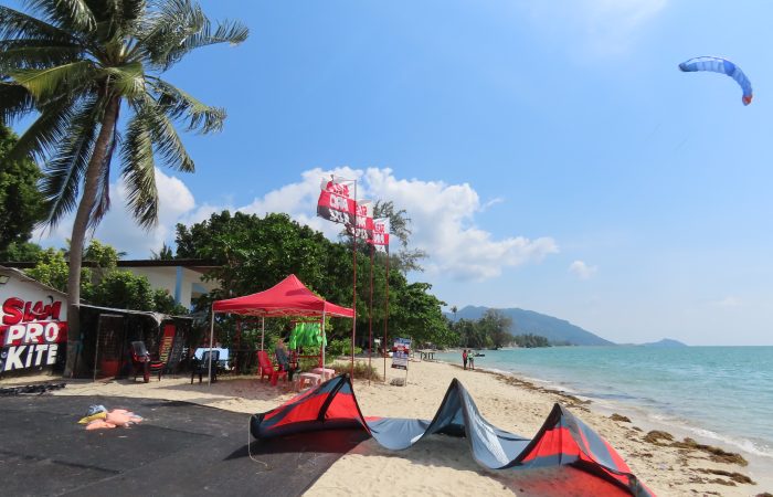 Siam Pro Kite kitesurfing center at Baan Tai Beach in Koh Phangan with a kite parked on the sand and another kite flying above the tropical beach on a sunny windy day.