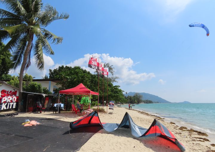 Siam Pro Kite kitesurfing center at Baan Tai Beach in Koh Phangan with a kite parked on the sand and another kite flying above the tropical beach on a sunny windy day.