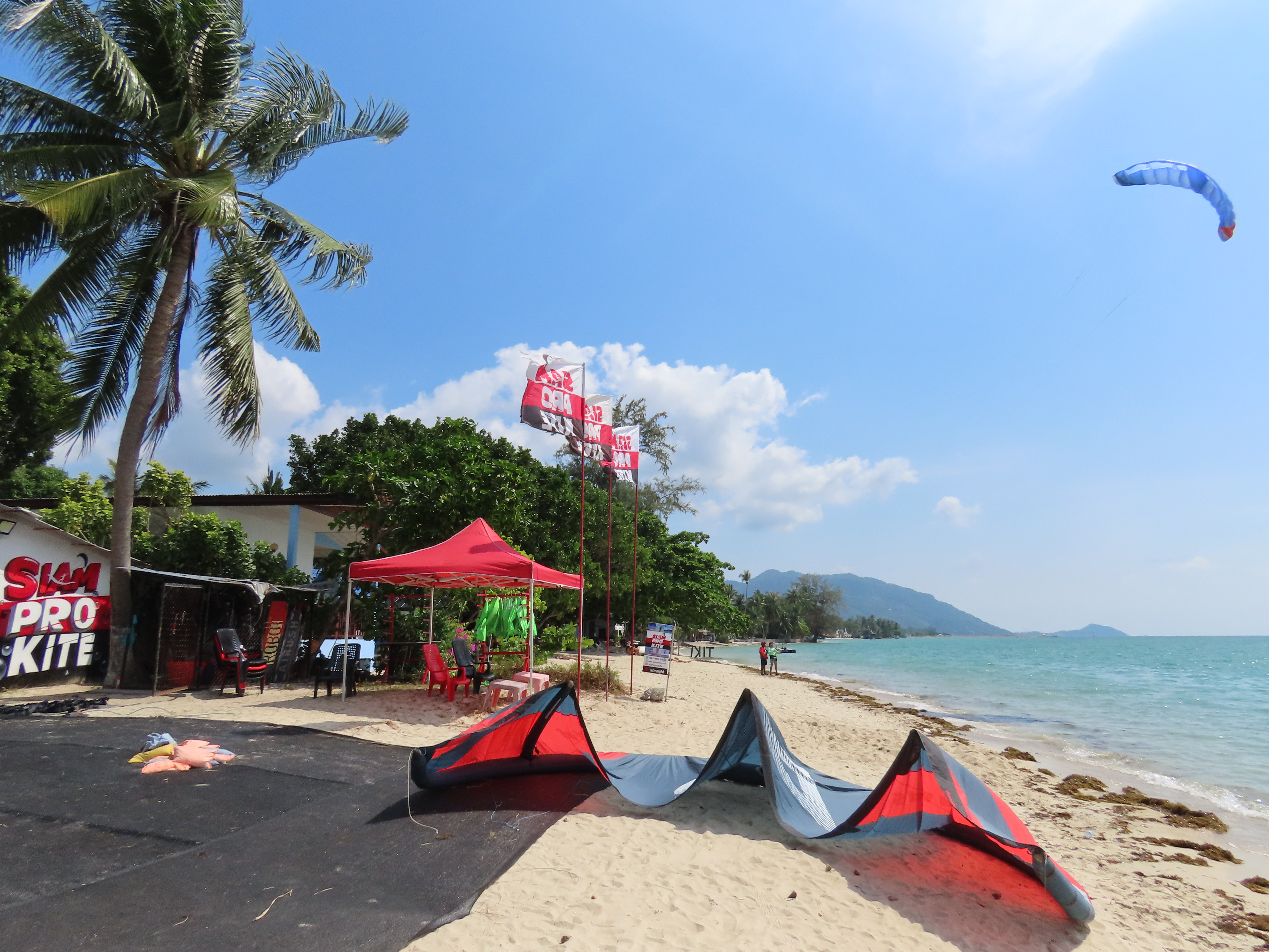 Siam Pro Kite kitesurfing center at Baan Tai Beach in Koh Phangan with a kite parked on the sand and another kite flying above the tropical beach on a sunny windy day.