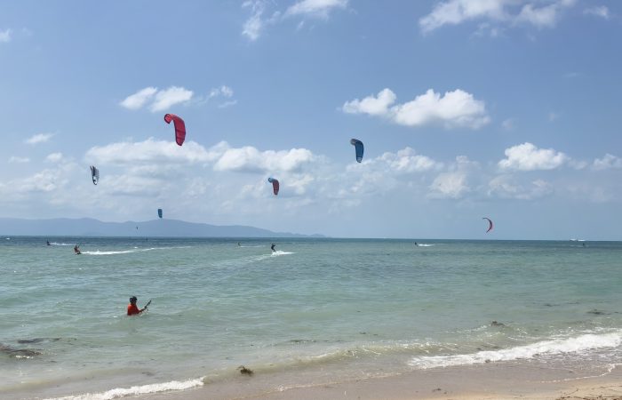 Many kitesurfers riding and launching kites on a very windy day at Baan Tai Beach Siam Pro Kite Koh Phangan Thailand with multiple colorful kites filling the sky and active riders on the water.